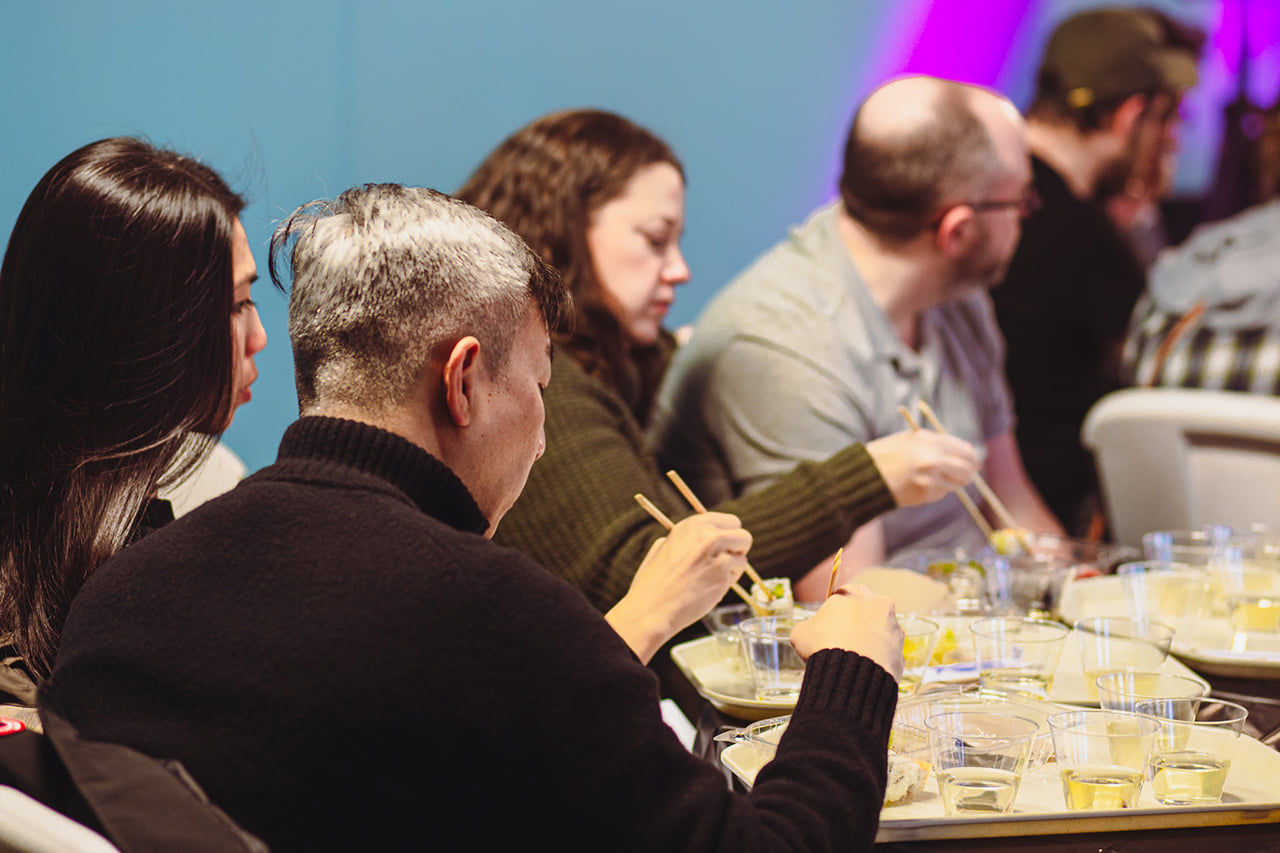 Several people seated at round tables, eating sushi and drinking sake with a blue wall in the background